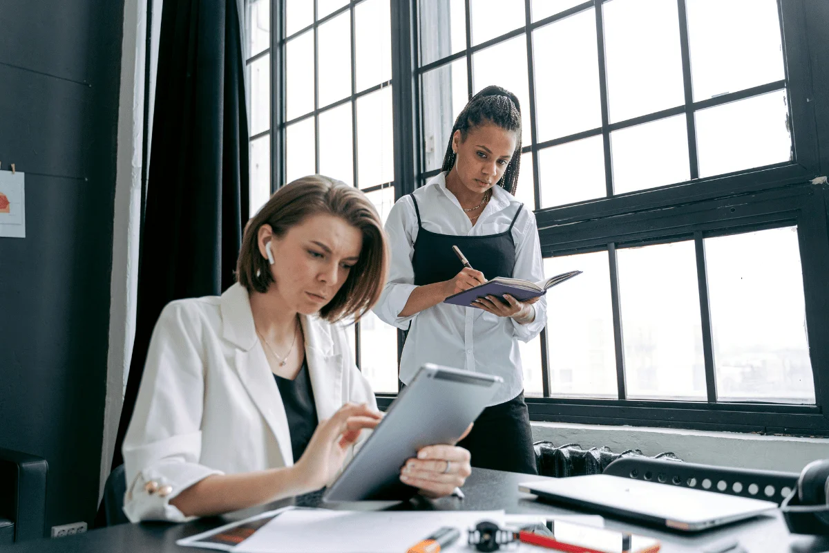 Two women work in an office. One sits using a tablet, focused, while the other stands writing in a notebook.