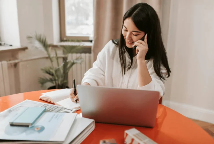 Confident woman writing in a notebook while managing a phone conversation.
