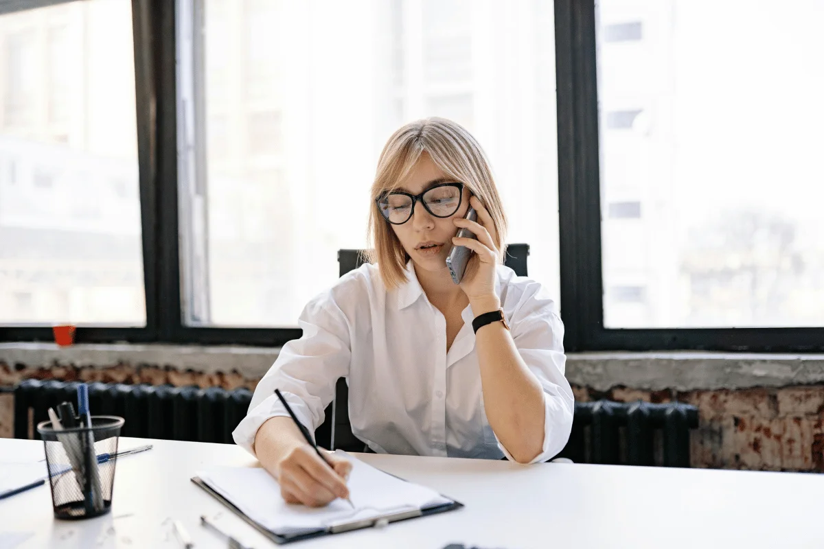 Businesswoman talking on phone while taking notes at desk.