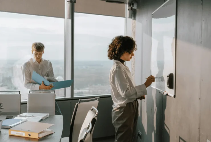 Two people in a bright office. One writes on a whiteboard, focused, while the other reviews a document by a window with a city view.