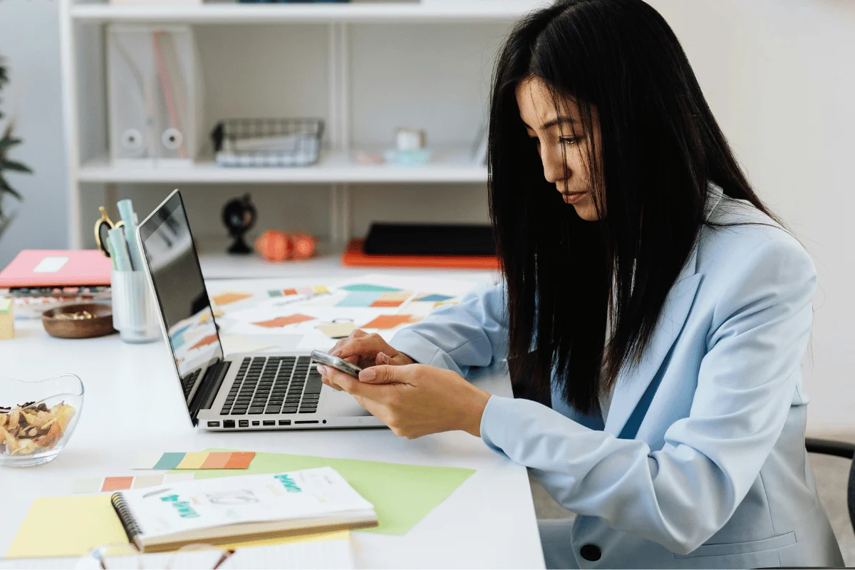 A woman working in an office, using a smartphone while sitting in front of a laptop, surrounded by colorful stationery and design materials.
