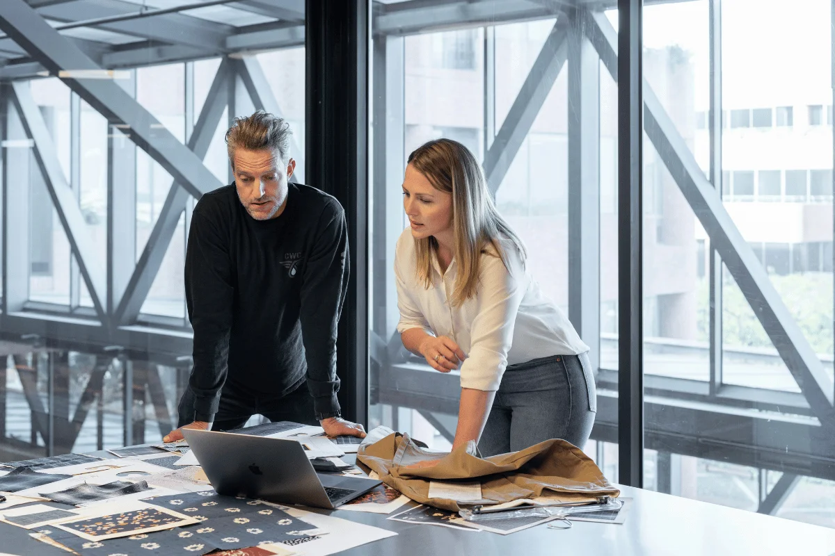 A man and woman lean over a table in a modern office, studying documents and a laptop. Large windows provide a bright, open atmosphere. Their expressions are focused and collaborative.