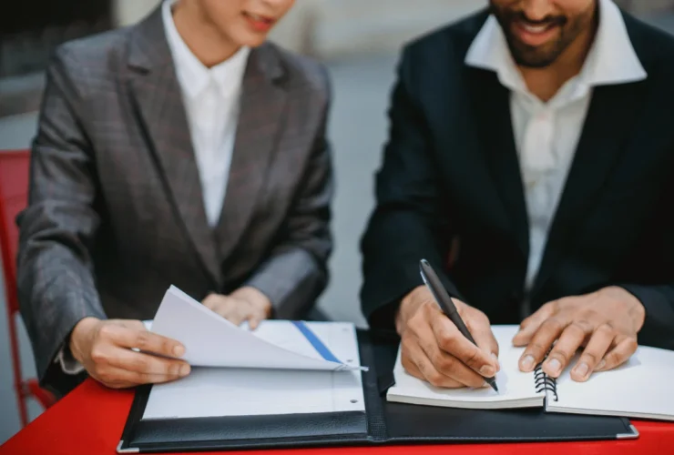Two people in business attire collaborate at a table, reviewing documents. One takes notes in a notebook, conveying professionalism and teamwork.