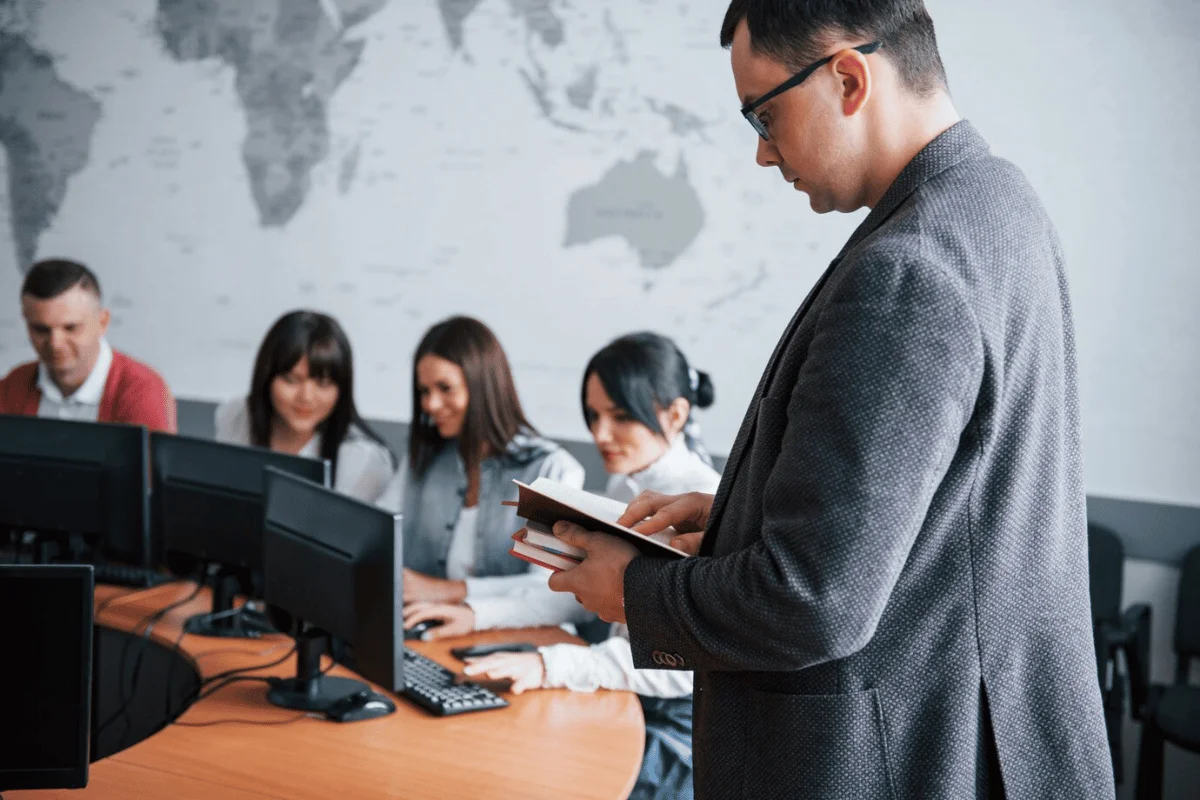 A man standing in front of a group of people in an office, holding a notebook while they work on computers.