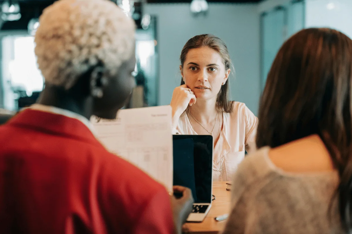 Two women engaged in conversation at a table with a laptop and documents, set in a modern cafe environment.