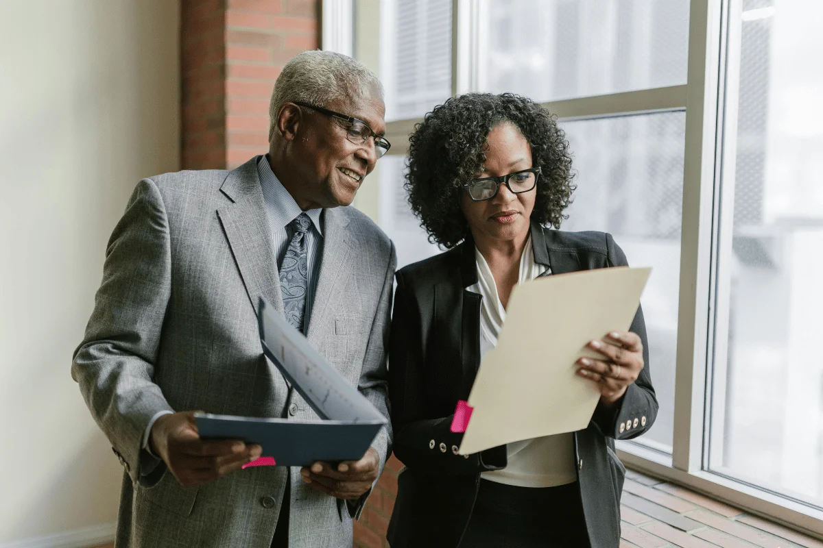 A man in a gray suit and a woman in a black blazer are discussing documents next to a large window in a well-lit office.