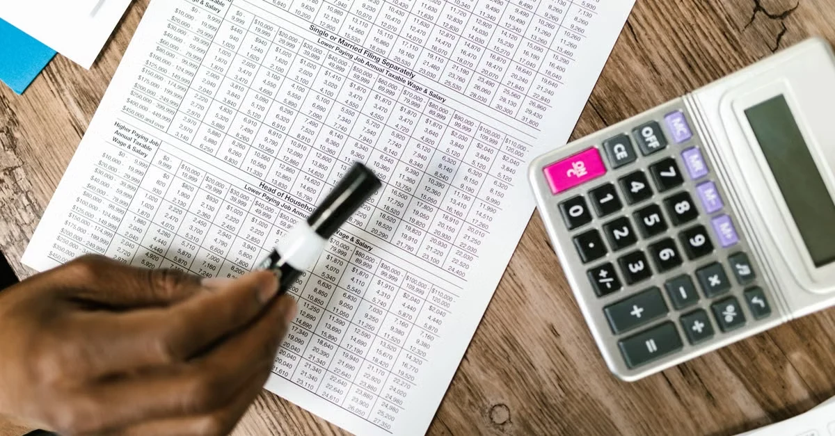 Person analyzing financial spreadsheet with calculator for remote EA cost breakdown on wooden desk.