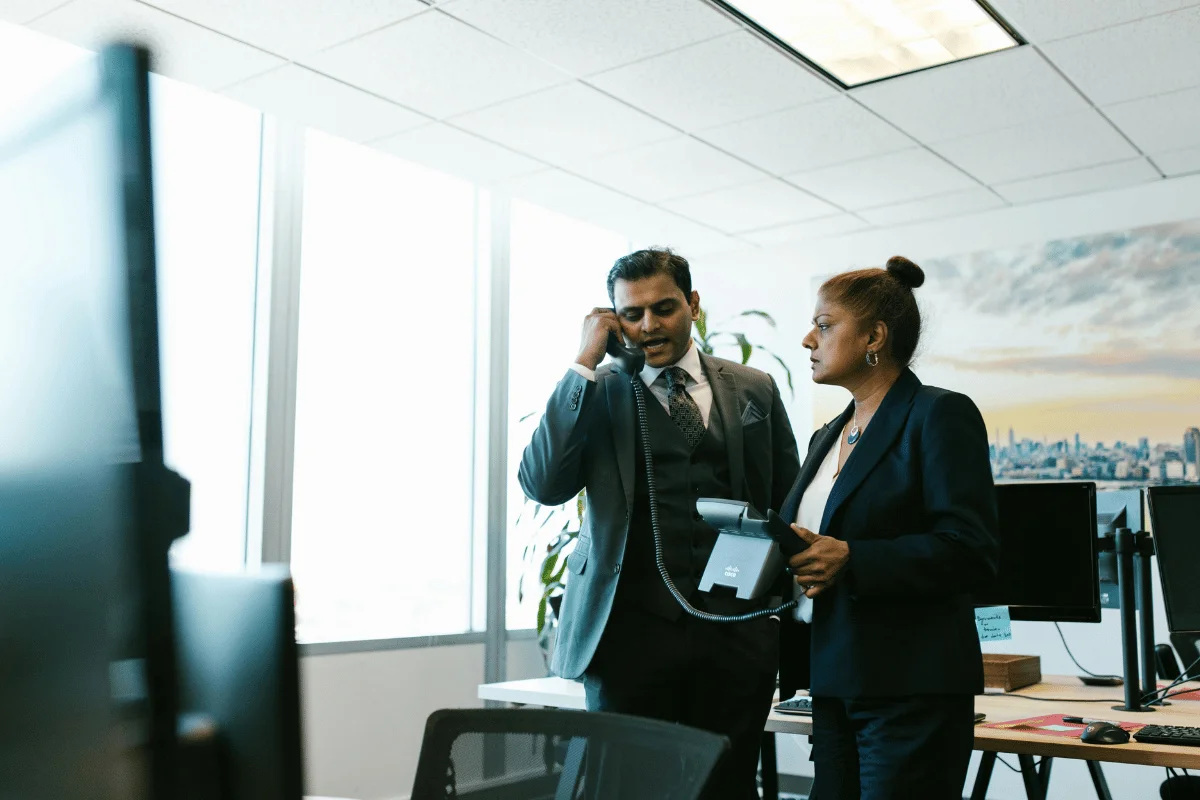 Businessman on office phone while colleague listens beside him.