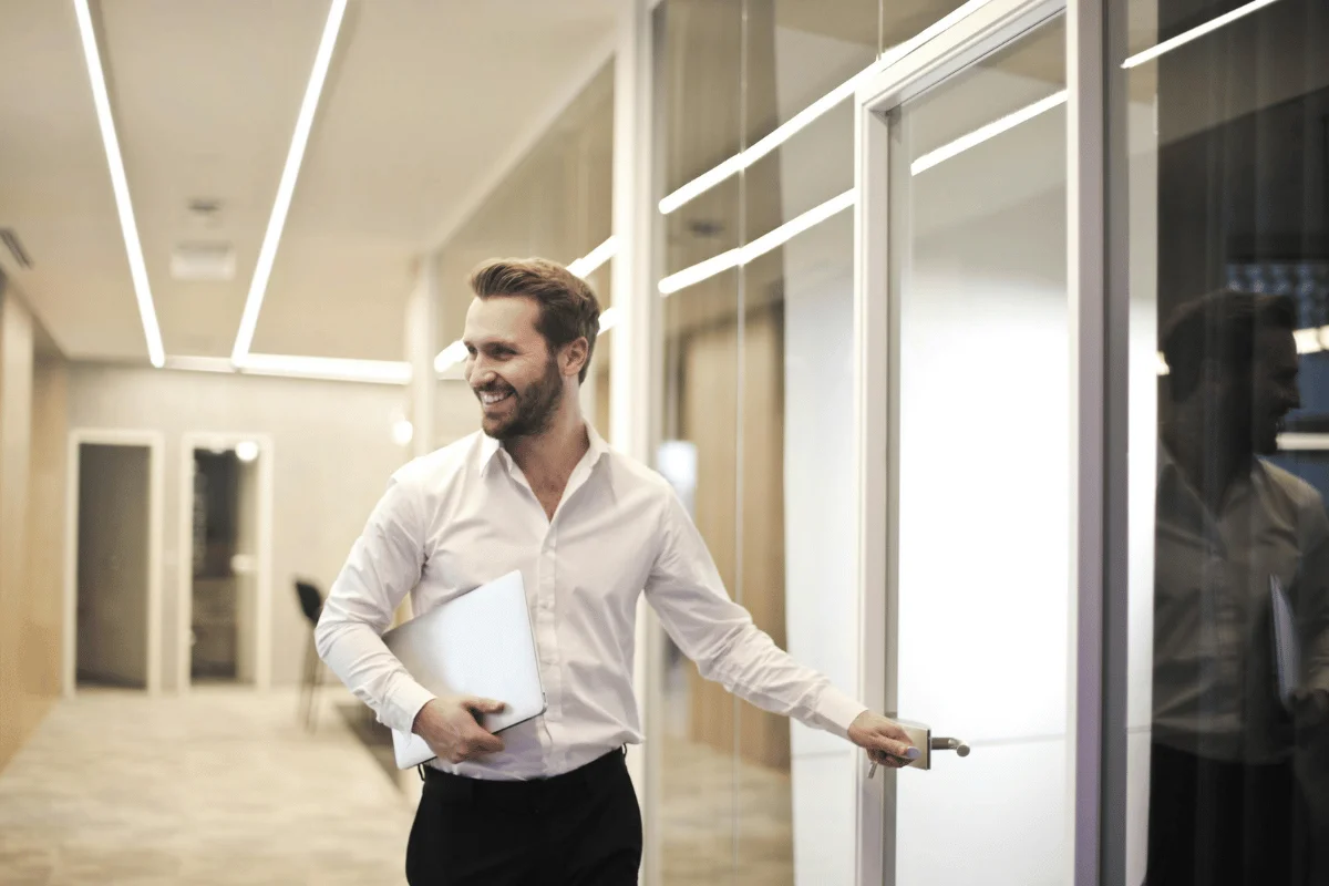 Smiling businessman opening office door with laptop in hand.