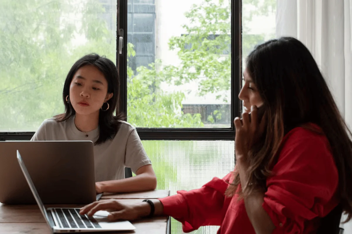 Two women working together at a desk with laptops by a window.