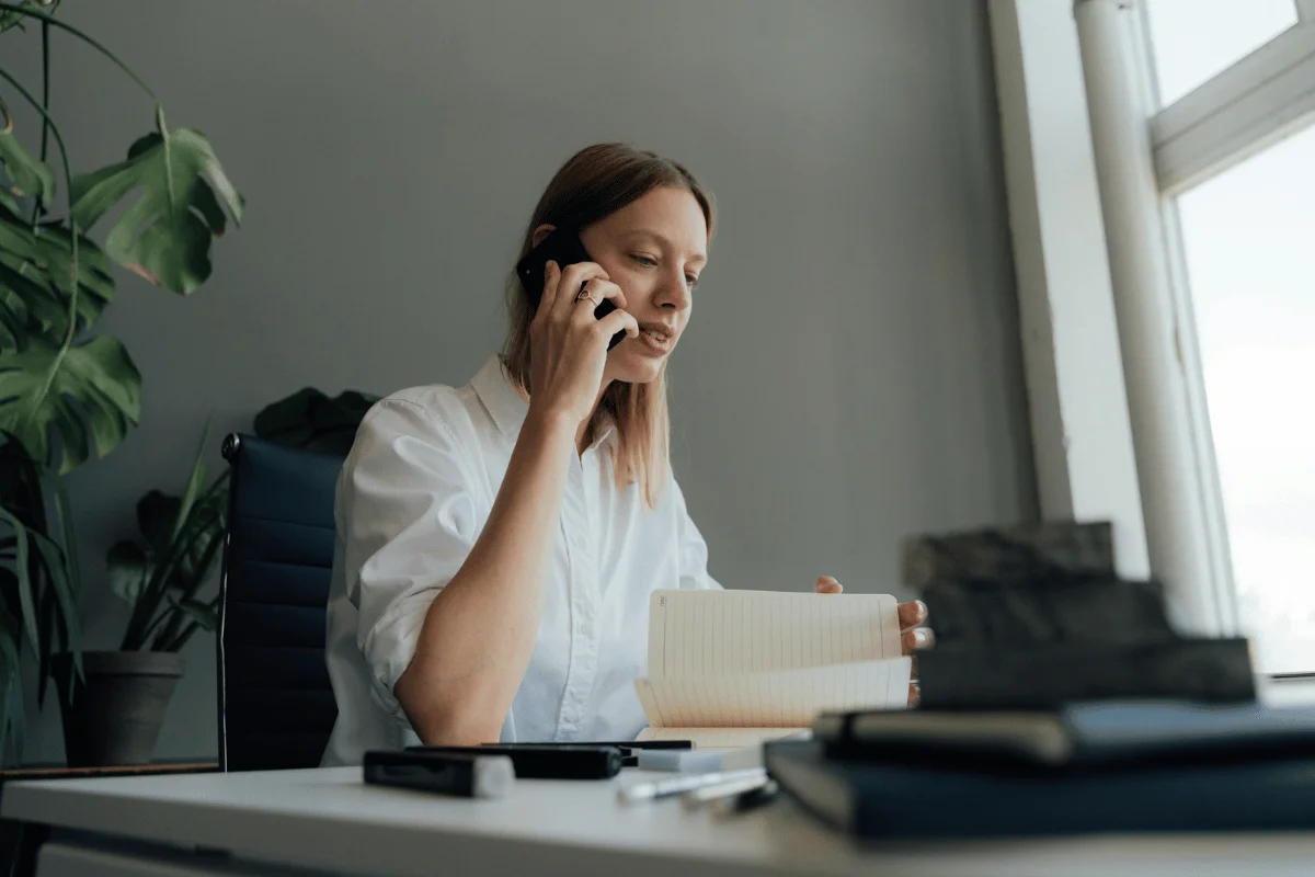 A person in a white shirt holds a notebook while talking on the phone at a desk with stationery and a plant in the background.