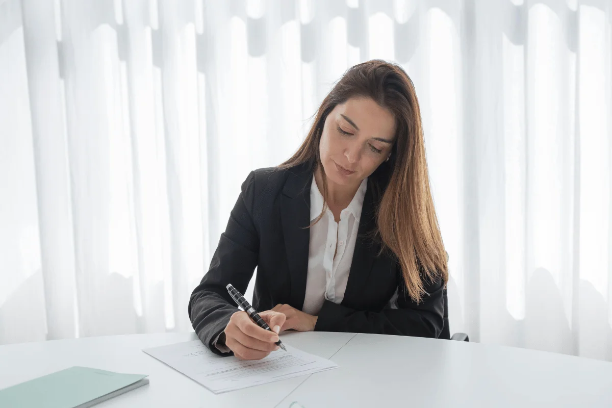 A woman in a business suit writes at a white desk with a focused expression, illuminated by soft light through sheer curtains in the background.