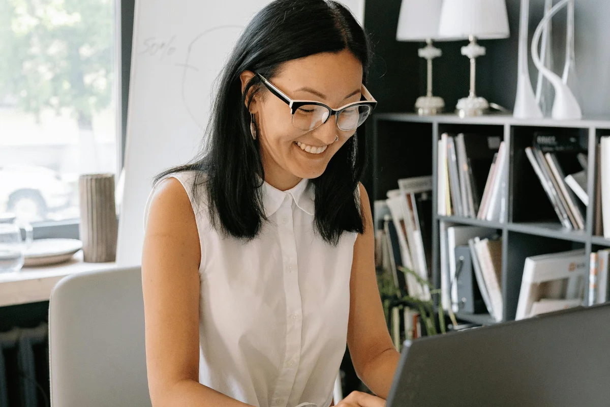 A woman in a white shirt sits at a desk, working on a laptop in a modern office with bookshelves and decorative lamps.