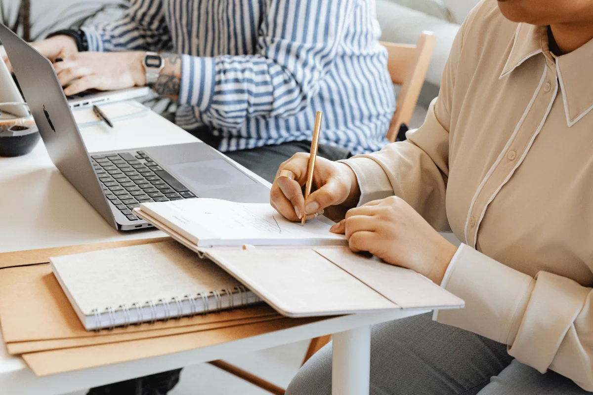 A person writes notes in a notebook while another uses a laptop, surrounded by stationery and a calm workspace.