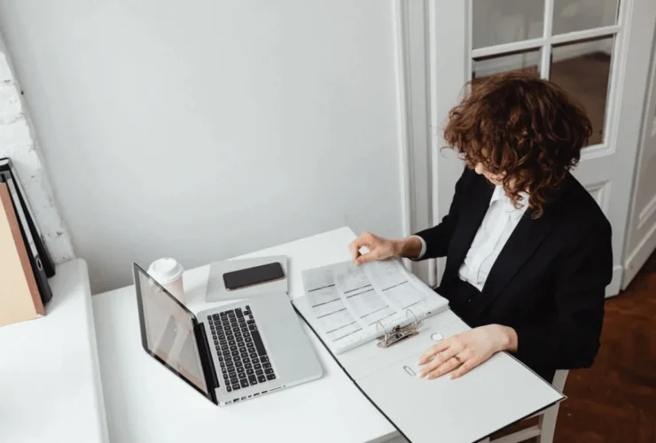 A woman in a black blazer reviews documents in a binder at a white desk with a laptop, smartphone, and coffee cup nearby.