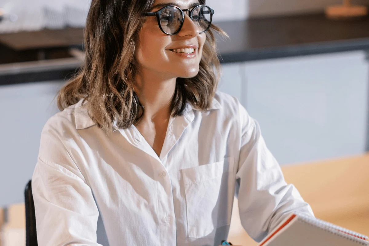 A woman with shoulder-length hair and glasses smiles while seated at a table. She wears a white shirt, conveying a cheerful and professional tone.
