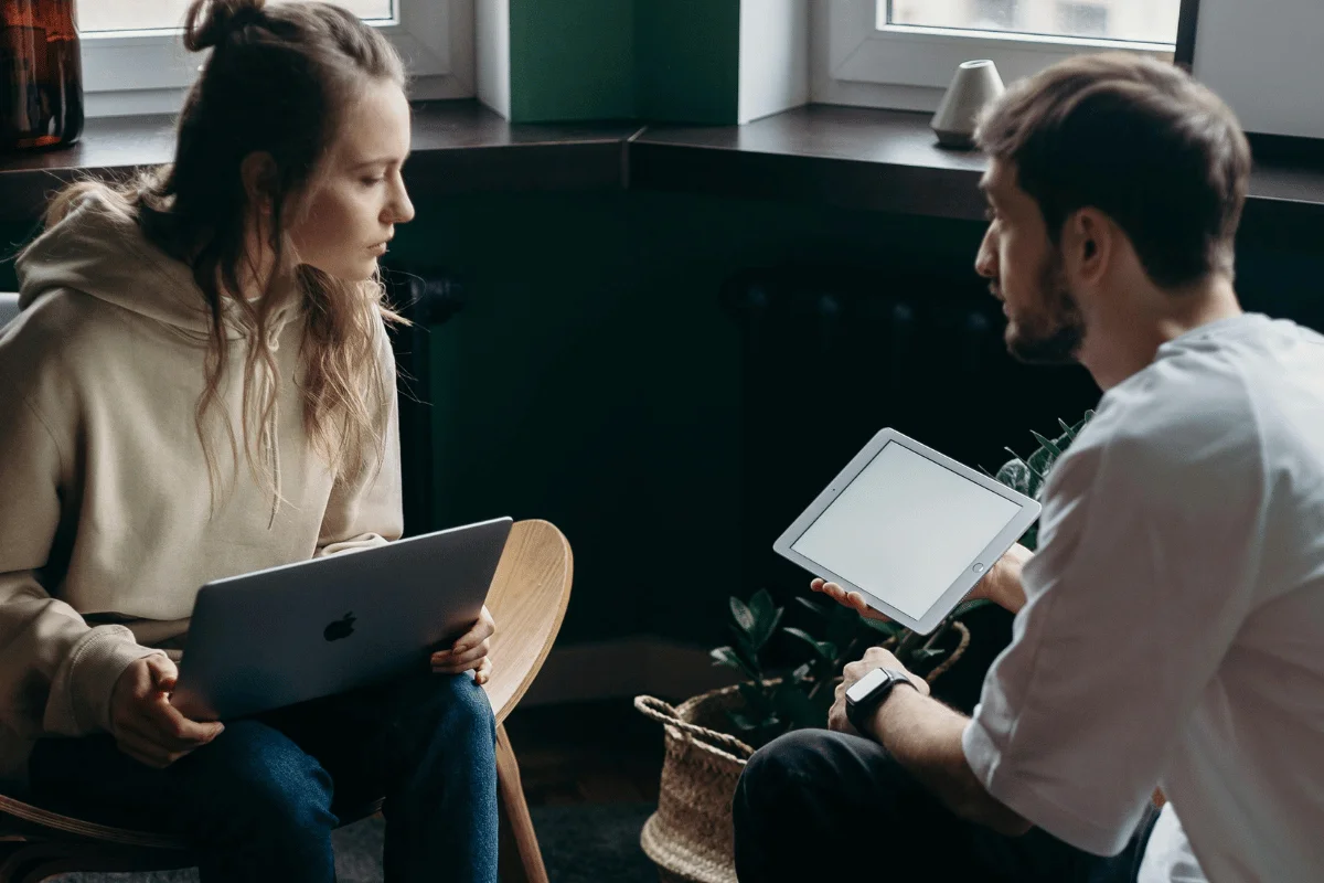 Man with a smartwatch presenting ideas on a tablet to a woman with a laptop.
