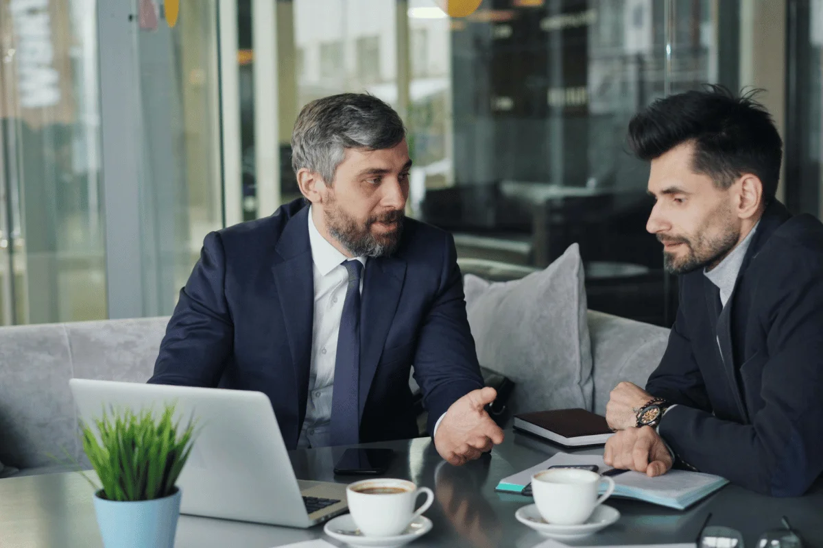 Two businessmen in suits discussing work over coffee at a modern office café table.