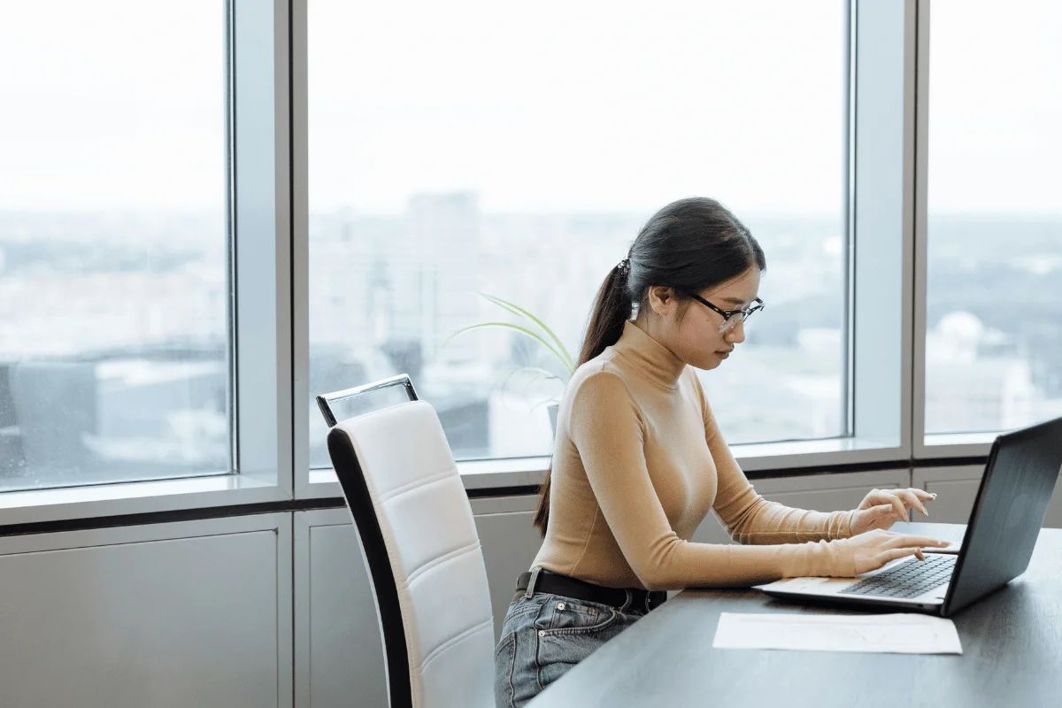 Professional assistant focused on typing at laptop near large windows.
