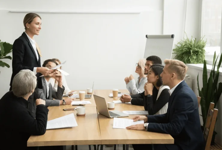 A professional woman stands, speaking to a group seated at a conference table.