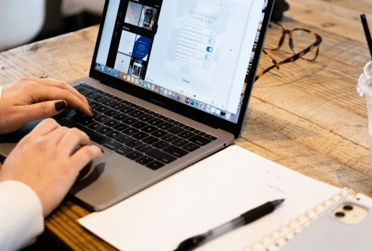 A person types on a MacBook Pro, displaying a presentation about hiring a virtual assistant, with a notebook and pen nearby.