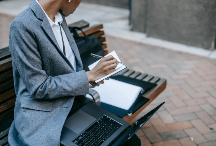 Person typing on a laptop while holding a pen above an open notebook with sticky notes.