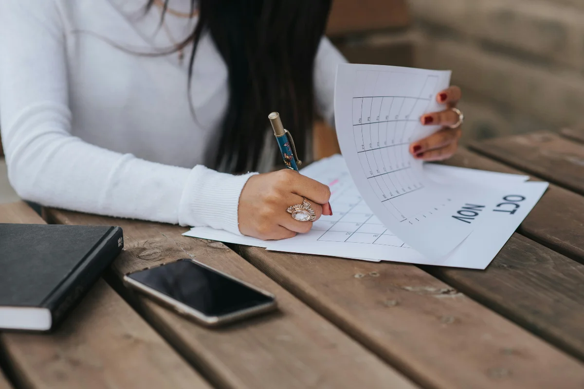 A person writes on a planner at a wooden table, with a smartphone and a notebook nearby, showing focus on organizing tasks.