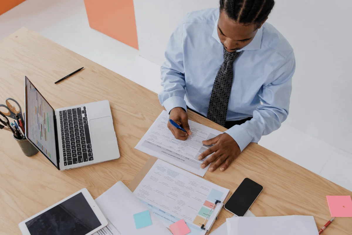 Man working on paperwork with a pen, laptop, and tablet on his desk, surrounded by sticky notes and a phone.