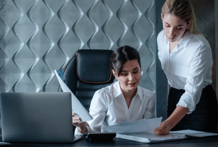 Two businesswomen in white shirts review documents together at an office desk with a laptop and calculator.