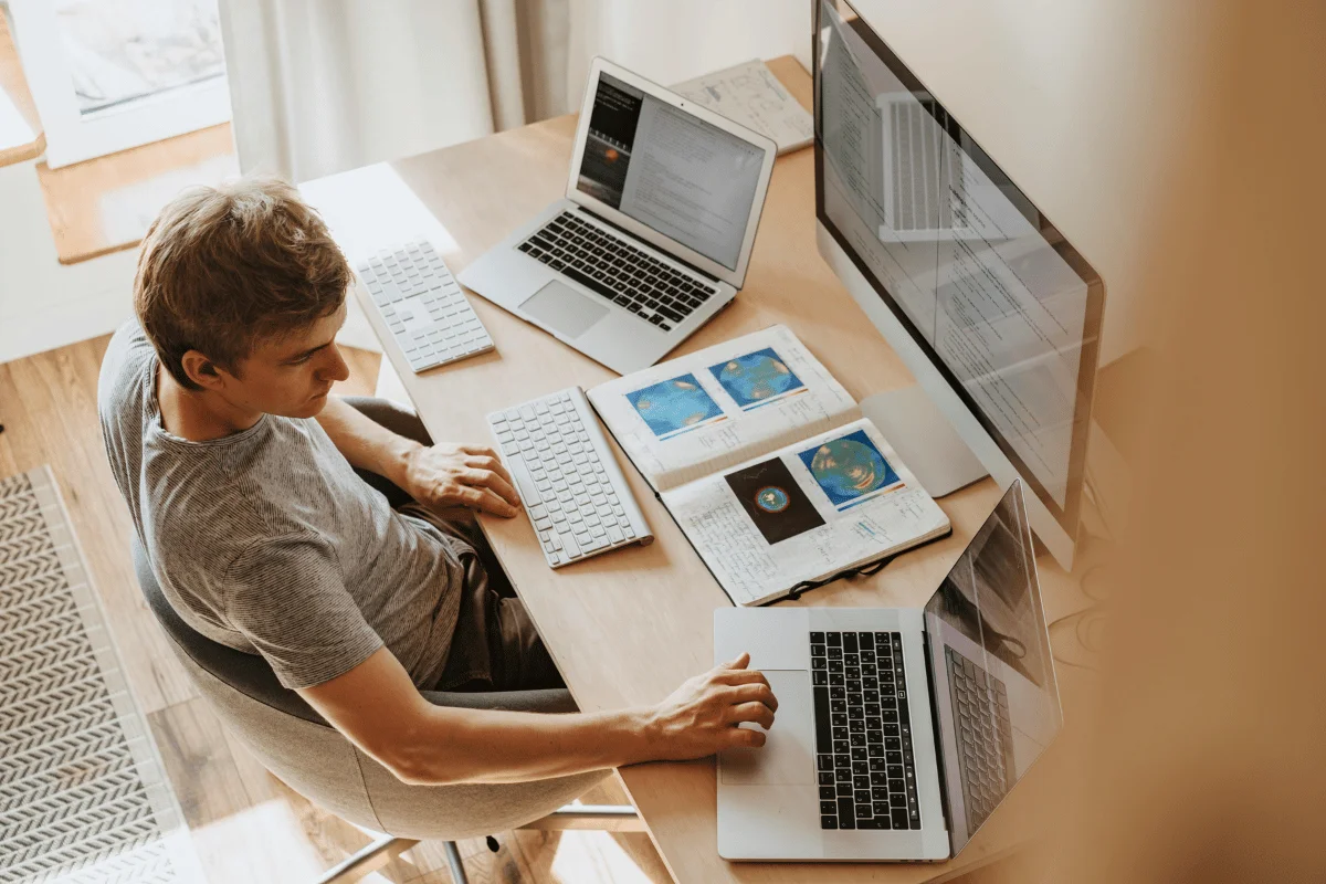 A person sits at a wooden desk working on two laptops and a large monitor, with an open notebook displaying colorful circular diagrams in the center.