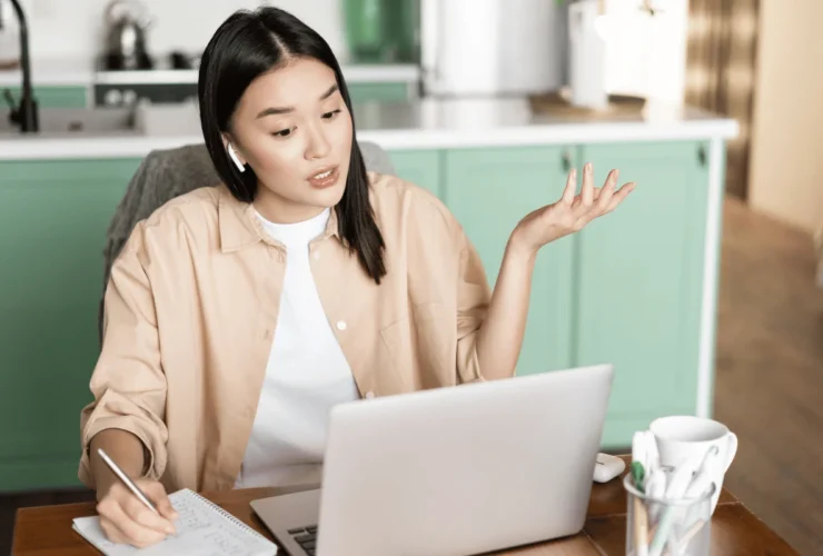 Person with earbuds gestures with one hand while writing notes at a table with an open laptop in a home kitchen.
