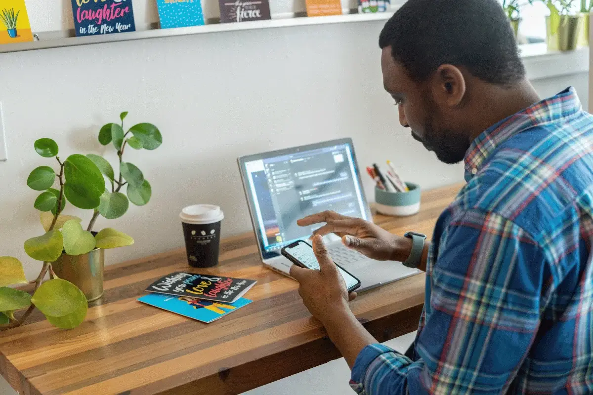 Man using a smartphone while working on a laptop at a home office desk with plants, coffee, and inspirational cards for productivity.