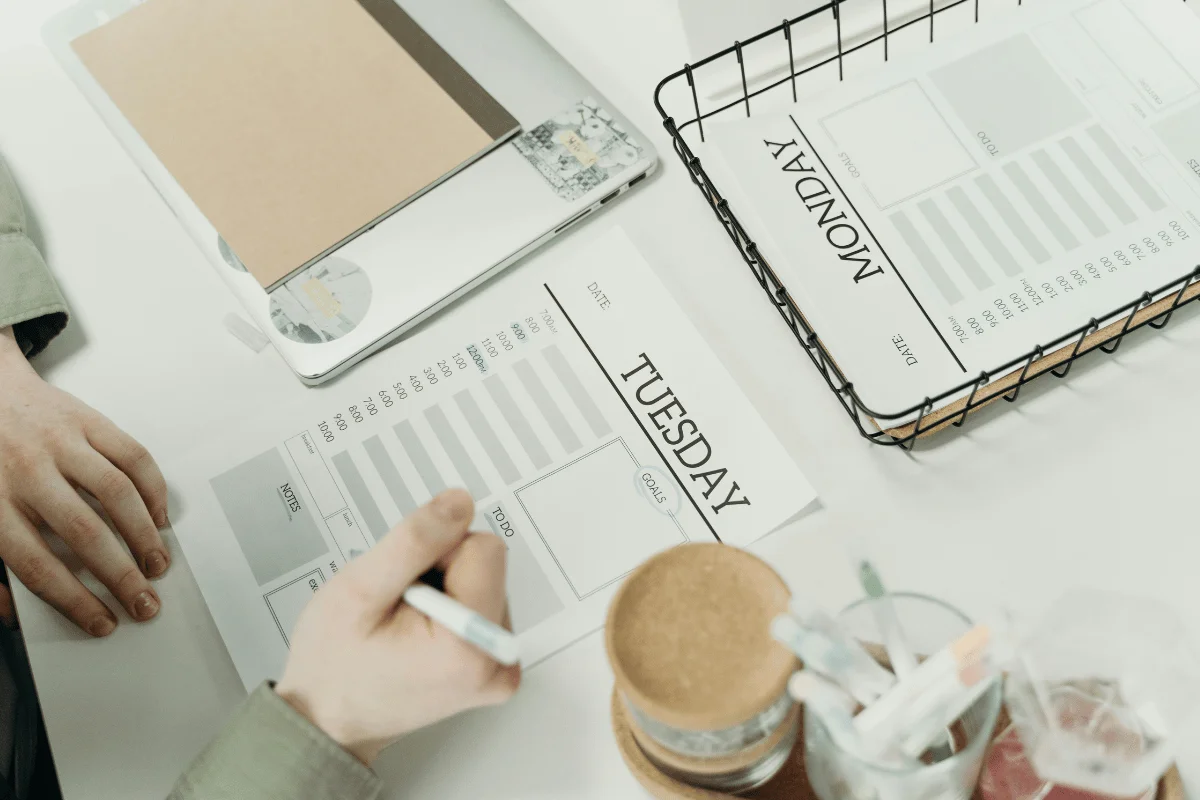 A person writes on a "Tuesday" planner beside a "Monday" planner in a wire basket.