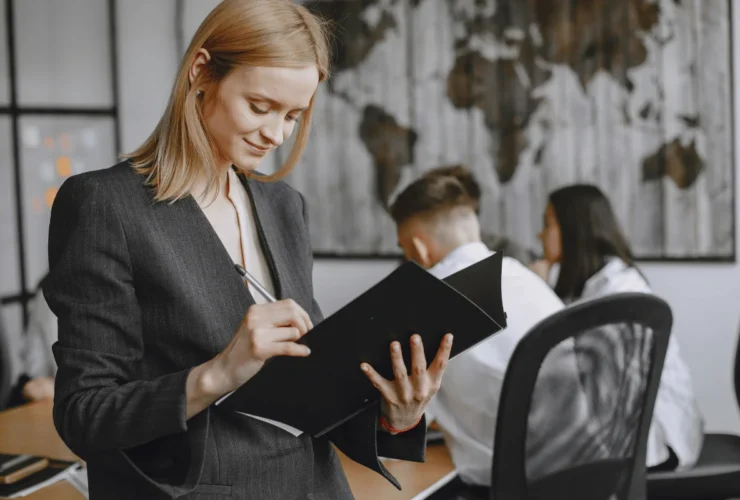 Professional woman in a suit smiling while writing in a folder in a modern office, with colleagues working near a world map in the background.