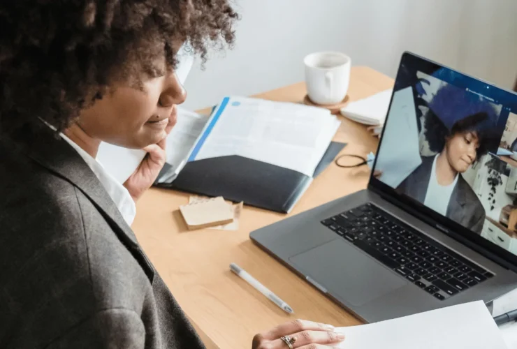 A woman in a blazer participates in a video call on a laptop at a wooden desk, surrounded by papers and a coffee cup, conveying a professional setting.