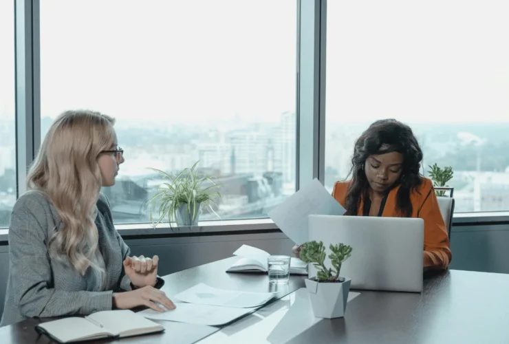 Two women in a modern office, one with blonde hair talking, the other working on a laptop