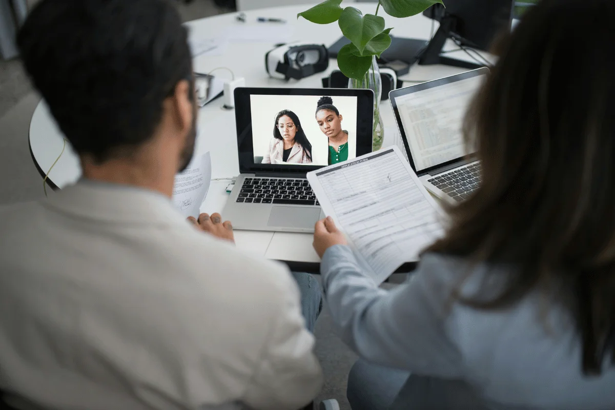 Virtual team collaboration with one person holding papers while viewing a video conference on a laptop.