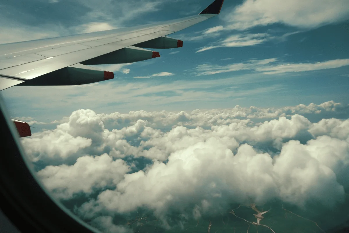Airplane wing seen from a passenger window above thick white clouds and blue sky.