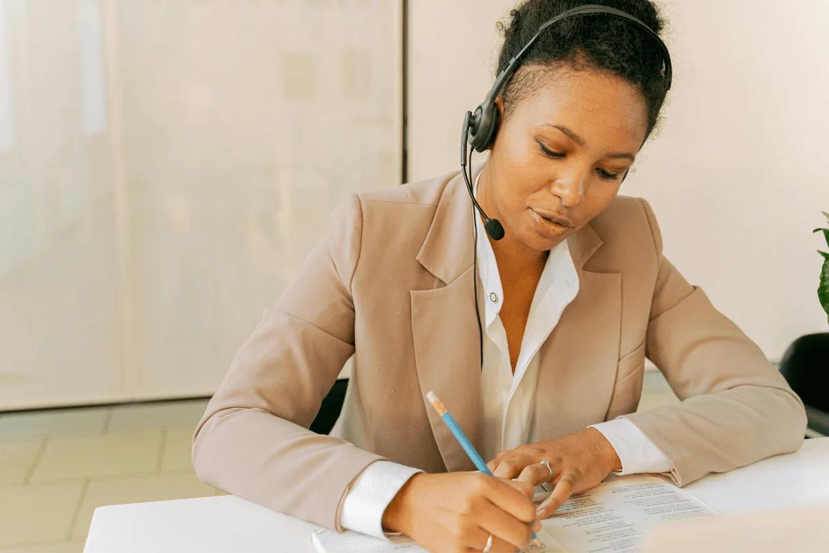 A professional woman in a beige blazer wearing a headset, taking notes while working at a desk.