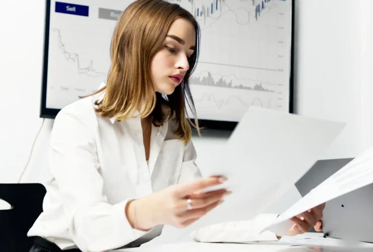 Professional woman reviewing documents at desk with financial charts displayed on monitor behind her.