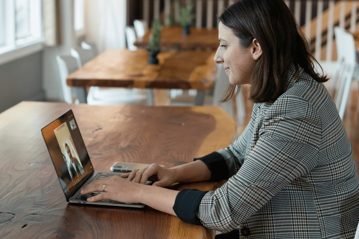 Woman using a laptop for a video call while seated at a large wooden table in a bright workspace.
