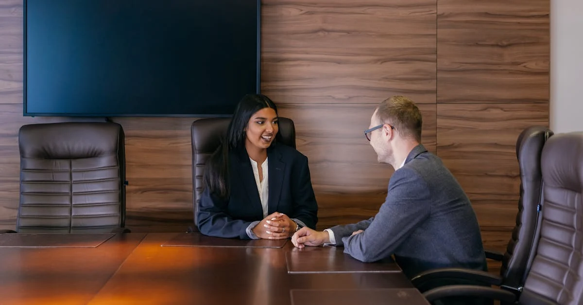 Two professionals in business attire discussing at conference table with TV screen, representing US based executive.
