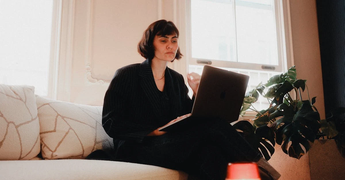Woman in black outfit working on laptop while sitting on couch near window and potted plant.