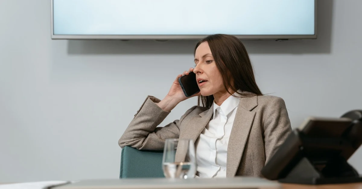 Executive assistant managing vendor relationship management on phone call at office desk with presentation screen behind.