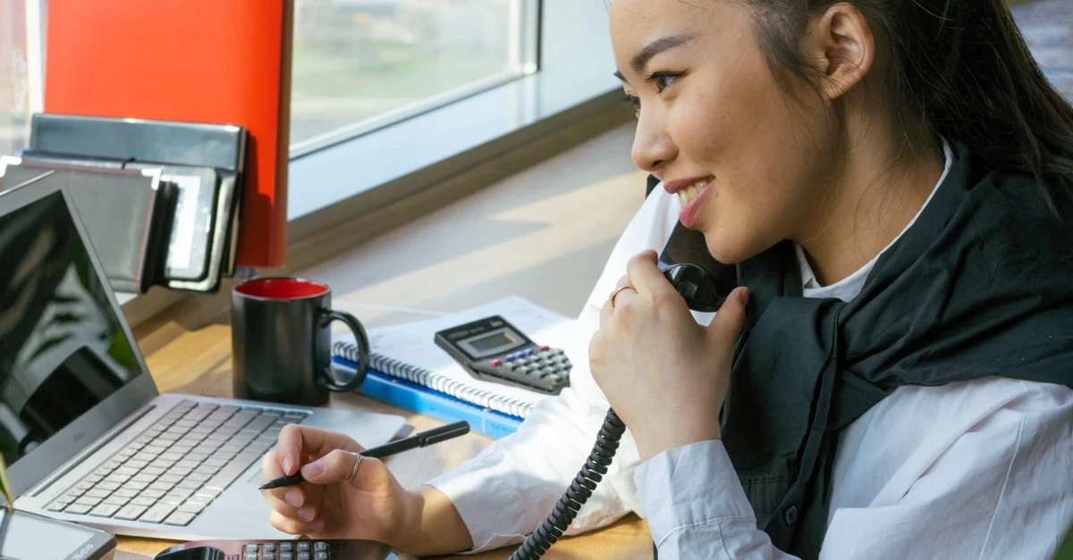 Professional woman on phone call at office desk with laptop and calculator for vendor relationship management discussion.