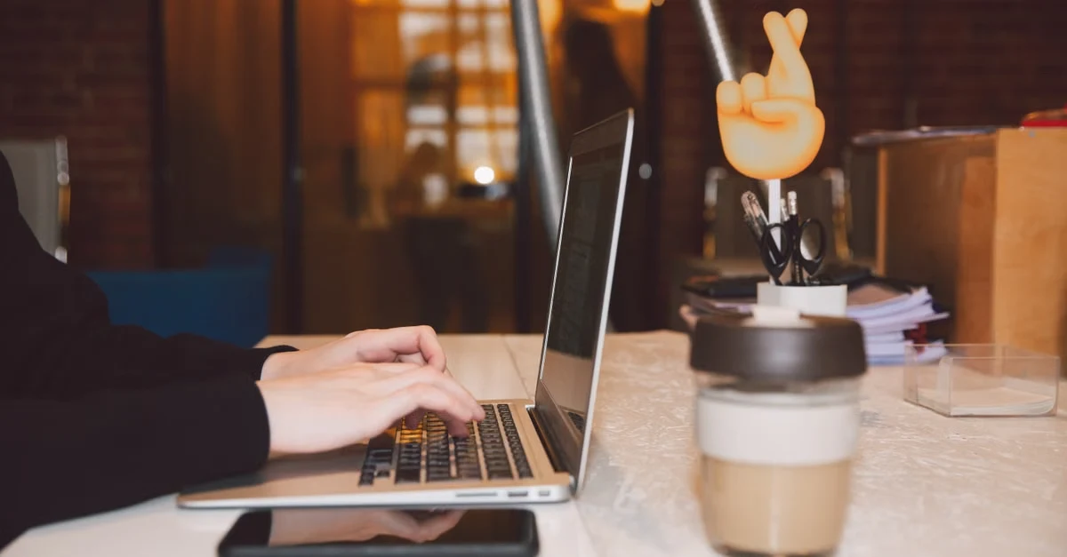 Person working on laptop at desk with coffee cup, phone, and desk lamp in evening workspace setup.