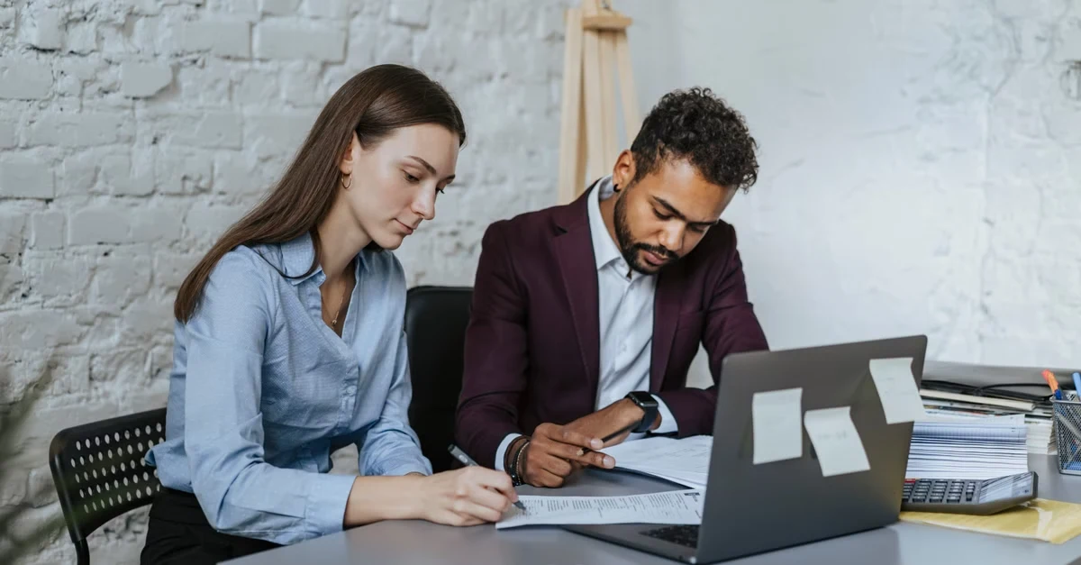 Executive assistants procurement professionals reviewing documents and laptop at desk in modern office workspace.