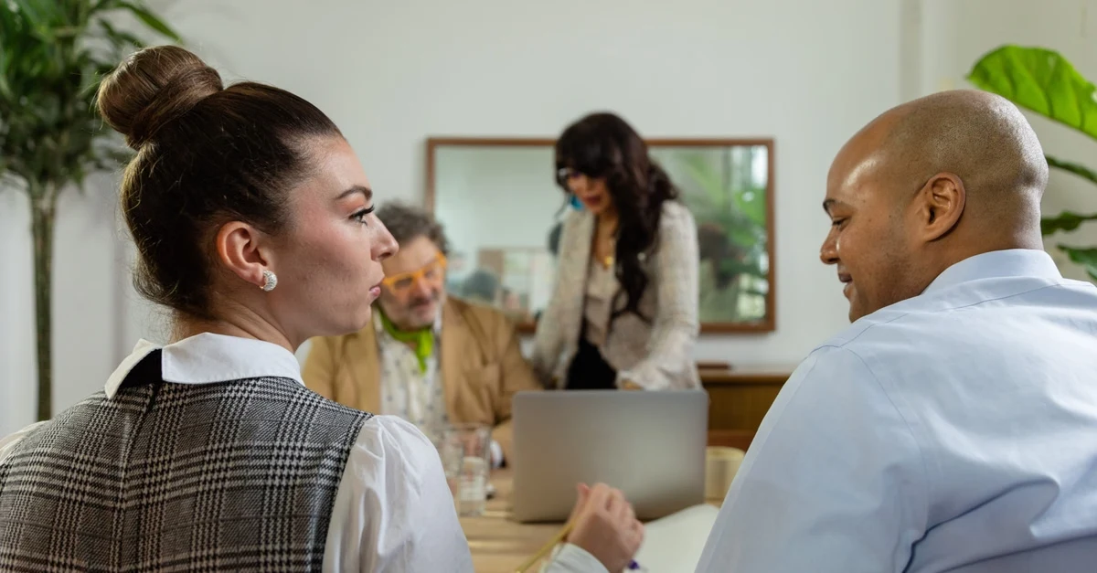 Two professionals discussing in modern office with plants and colleagues in background during business meeting.