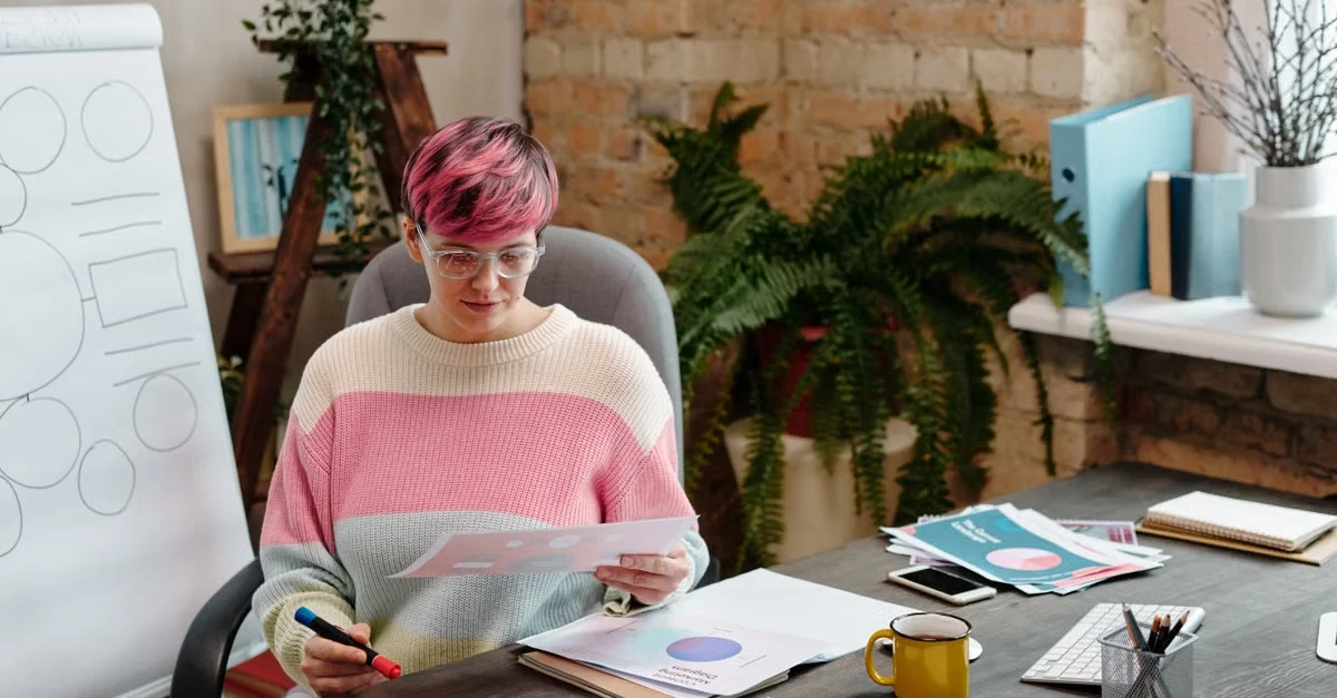 Executive assistant with pink hair reviewing keynote notes at desk with plants and whiteboard in background.
