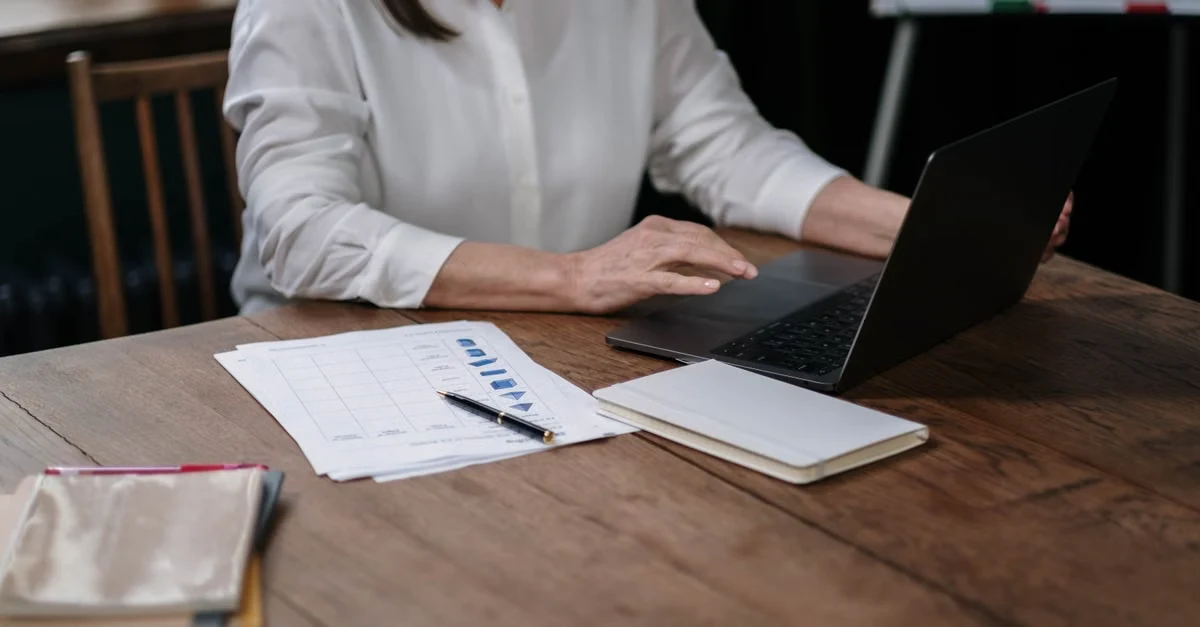 Professional woman working at wooden desk with laptop, documents, and notebook for remote business support companies.